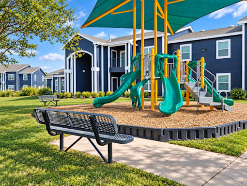 A playground with a green slide and a bench in front of a blue building.