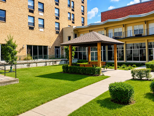 A gazebo is in the middle of a grassy area in front of a tan building.