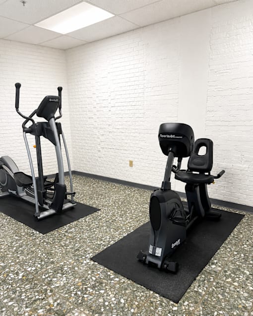 Two exercise bikes are placed on mats in a room with a white brick wall.