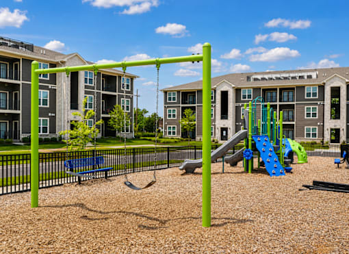 A playground with a green swing set and a blue slide in front of apartment buildings.