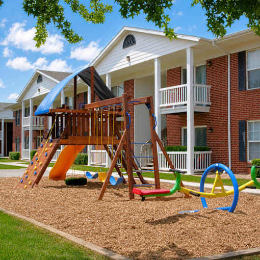 A playground with a slide and a wooden swing set in front of a brick apartment building.
