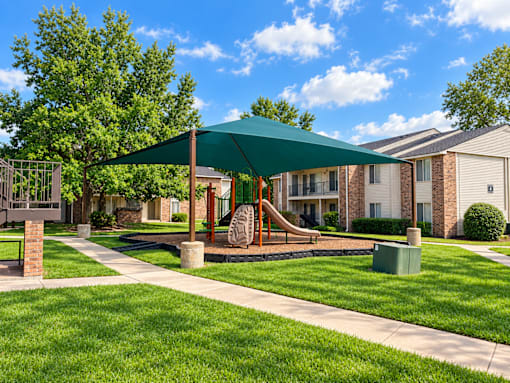 A playground with a green canopy and a slide is surrounded by a grassy area and apartment buildings.
