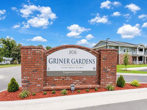 A sign for Griner Gardens Apartment Homes stands in front of a house.