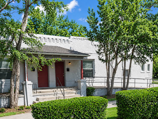 A white house with red doors and windows is surrounded by green bushes and trees.