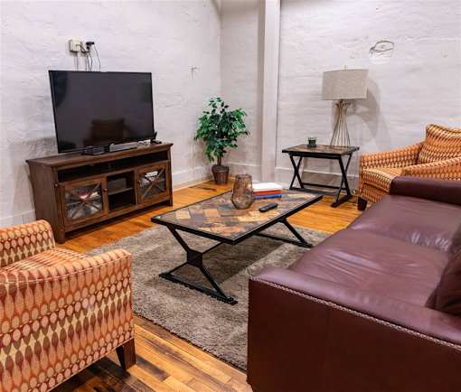 A living room with a brown leather couch, a patterned chair, a wooden cabinet, a glass coffee table, and a television.