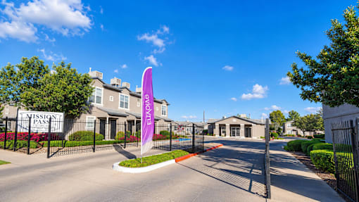 The image shows a gated entrance to a residential area named "Mission Pass" with a purple flag in the foreground.