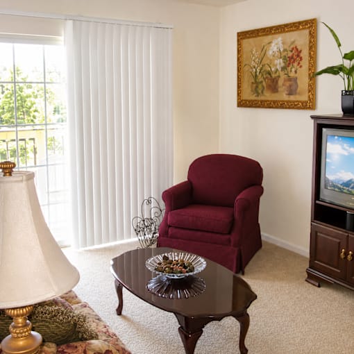 A red chair sits in a living room with a lamp and a painting on the wall.