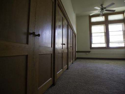 A room with a carpeted floor, wooden cupboards, and a window with a fan.
