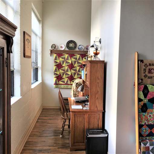 A room with a wooden desk and chair, a quilt hanging on the wall, and a window with blinds.