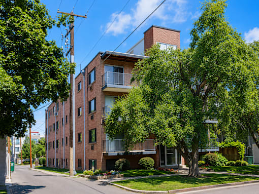 A red brick building with a balcony on the second floor is surrounded by green trees.