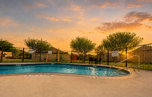 A pool surrounded by a fence with trees and a sunset in the background.