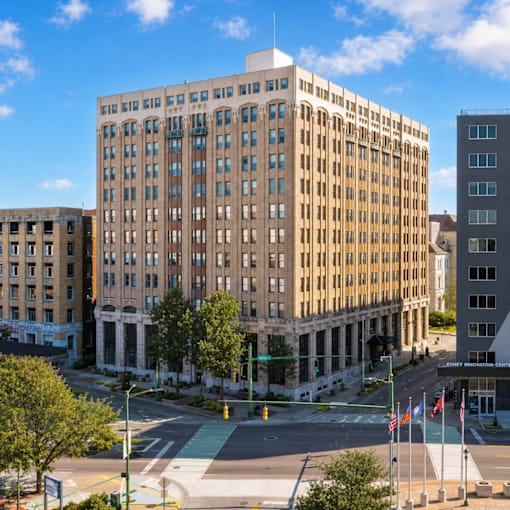 A large, beige building with many windows is in the foreground of a city street.