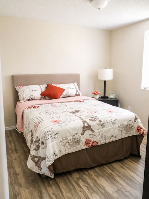 A bed with a white comforter and a pink pillowcase is in a room with a lamp and a picture frame on a nightstand.