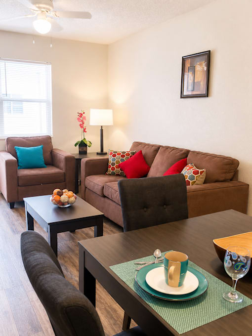 A living room with a brown couch, a brown chair, a brown armchair, a coffee table, a lamp, a vase with flowers, a bowl of fruit, a plate with a cup, and a picture on the wall
