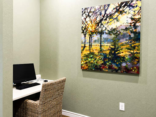 A painting of a forest scene is hanging on a wall above a desk with a computer.