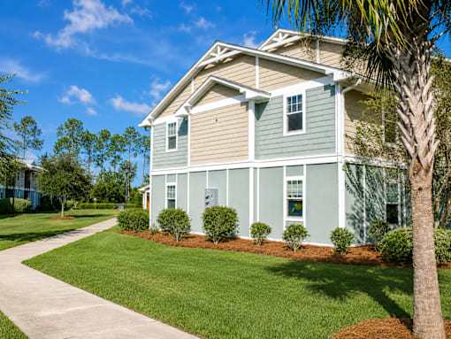 A house with a green and beige exterior is surrounded by a well-manicured lawn and a clear blue sky.
