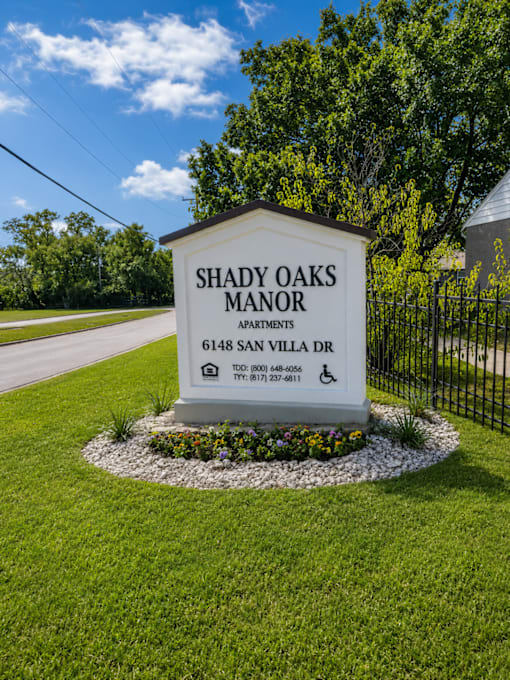 A sign for Shady Oaks Manor Apartments is displayed in front of a fence.
