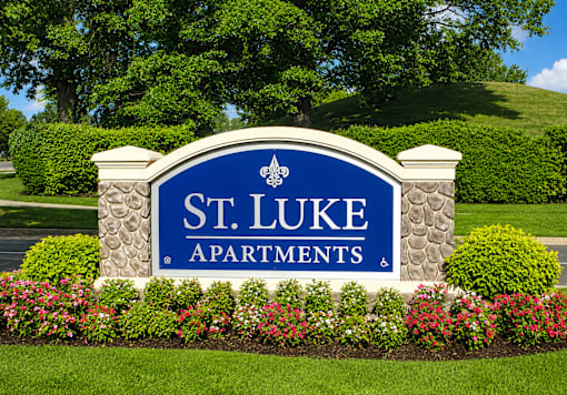 A sign for St. Luke Apartments is surrounded by flowers and greenery.