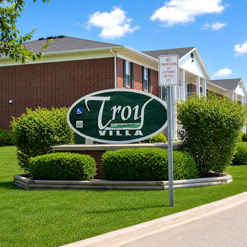 A sign for Troy Village is displayed in front of a brick building.