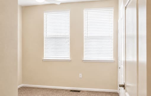 A room with a carpeted floor and two windows with blinds.