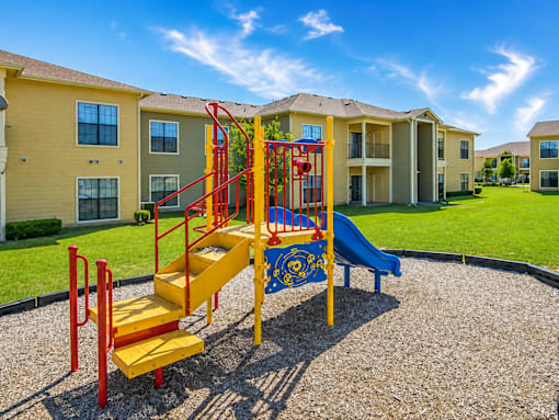 A playground with a yellow slide and red railings in front of apartment buildings.