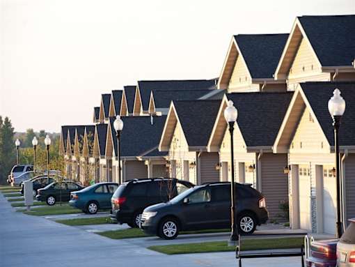 A street view of a residential area with cars parked on the side of the road and houses with triangular roofs.