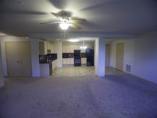 A spacious living room with a ceiling fan and a kitchen area in the background.