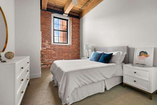 A bedroom with a white bed, dresser, and a window with a brick wall.