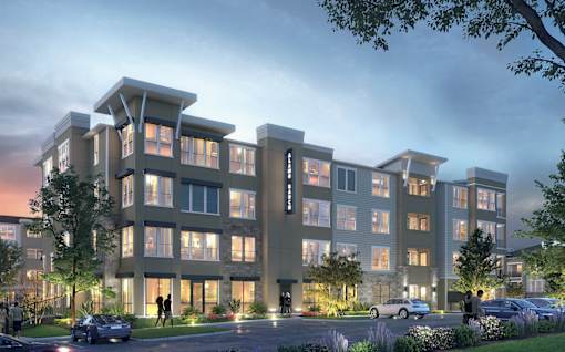 A modern apartment building is shown at dusk with lights on inside the building and cars parked in the driveway.