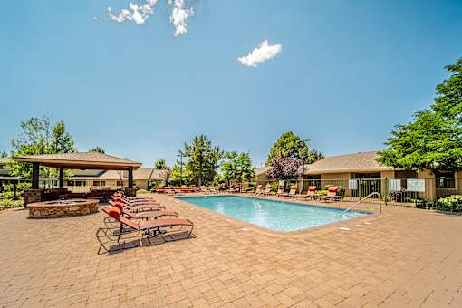 A pool surrounded by a brick patio with chairs and a building in the background.