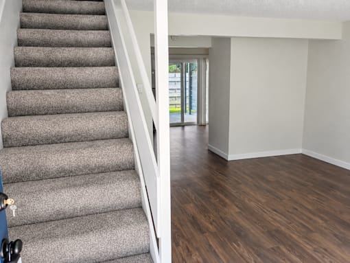 A staircase with a grey carpeted runner and white handrail leads to a bright room with a wooden floor and a glass door.