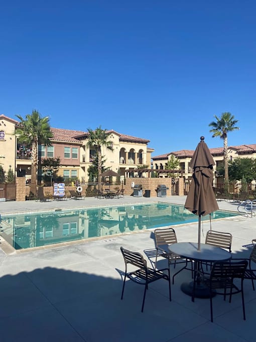 A table with chairs and an umbrella is in the foreground of a pool area.at The Villas 55+ II Hesperia, Hesperia  