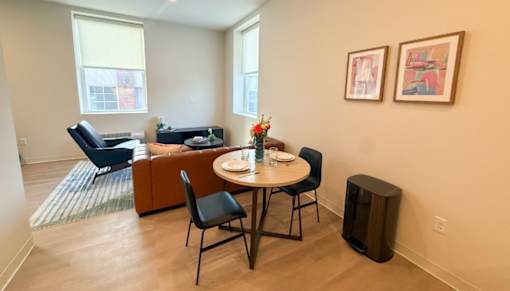 A living room with a brown couch, a blue chair, a small table with chairs, and two framed pictures on the wall.at The Jones Tower at the Collective, Pittsburgh, PA 15203