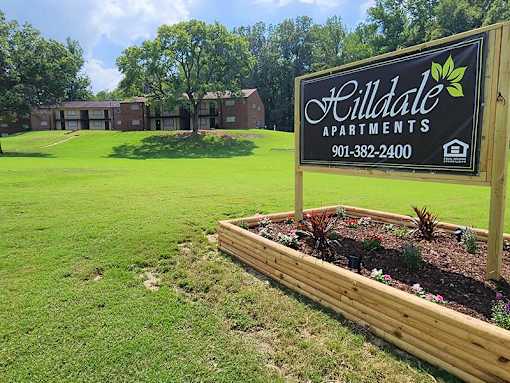 A sign for Hilldale Apartments stands in a flower bed in front of apartment buildings.