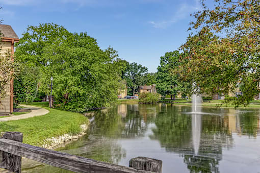 A serene park with a fountain in the middle of a pond surrounded by lush green trees.
