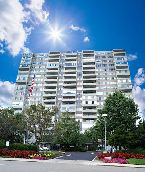 A tall building with an American flag on the side is surrounded by trees and flowers.