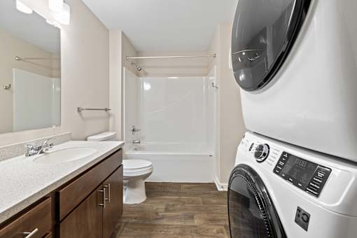 A white washing machine is in the foreground of a bathroom with a sink, toilet, and bathtub.
