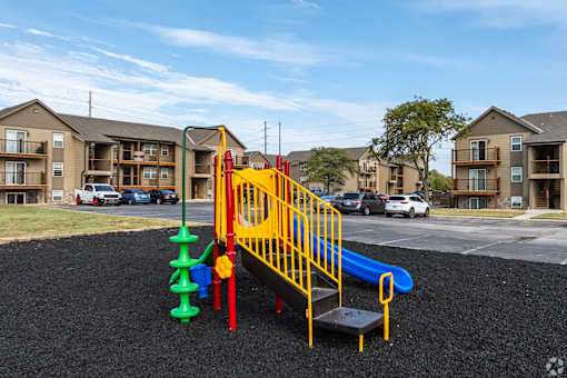 Playground with slide, swings, and seesaw, with apartment buildings in the background.