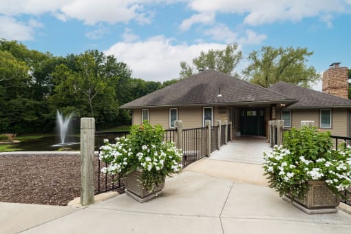 A charming Tanglewood building with a fountain on the left side and colorful flower beds in pots in the foreground. A bridgeway leads towards the building.
