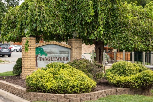 A welcoming Tanglewood sign marks the entrance to an apartment complex, surrounded by lush green bushes and framed by mature trees.