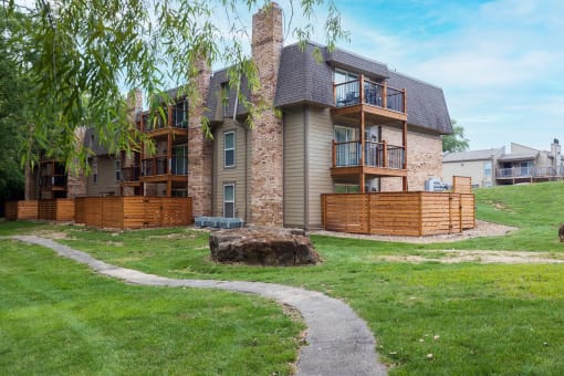 A view of the back of a house featuring a stone fireplace, a wooden balcony partition, a lush green yard, and a walkway.