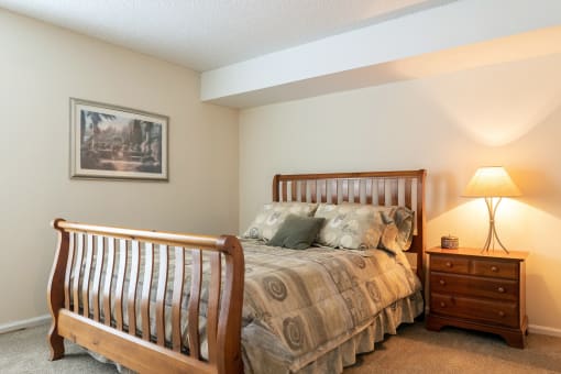 A cozy Tanglewood Model Unit Bedroom with beige walls features a comfortable bed, a nightstand with a lamp on the right side, and wall art adding a touch of personality to the left wall.