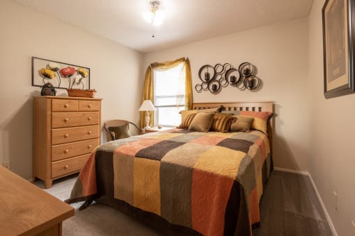 Cozy bedroom with a natural wood bed and dresser, beige walls, quilted bedding, a window bringing in natural light, and carpeted floors.