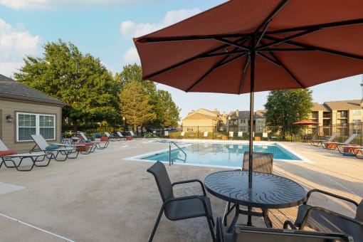 The pool area at The Lakes, featuring lounge chairs, a table with chairs, and an umbrella for shade.