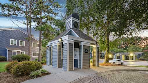 A small building with a steeple and a clock is surrounded by trees and houses.