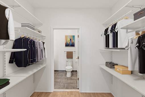 A white walk in closet with clothes hanging and a toilet in the background.