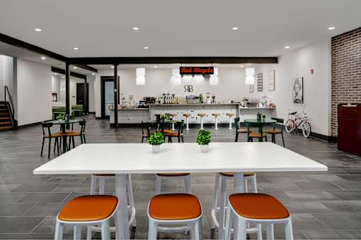A white table with orange chairs is in the foreground of a room with a bar in the background.