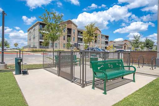 A green bench sits in front of a metal fence with apartment buildings in the background.