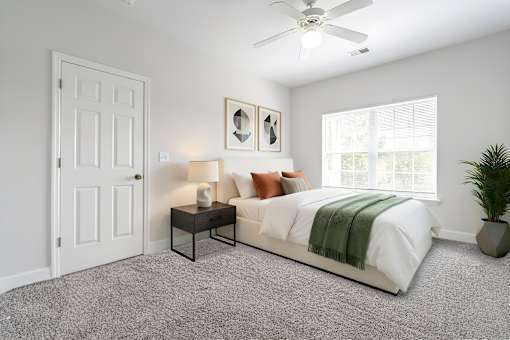 A bedroom with a bed, a nightstand, a lamp, a fan, and a plant.