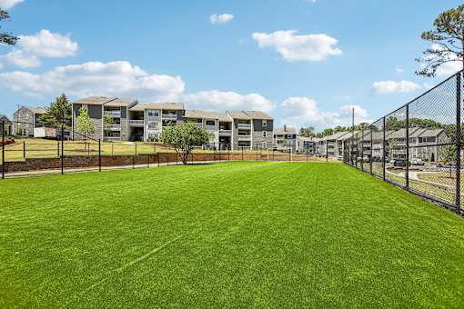 A large grassy field with a fence and apartment buildings in the background.